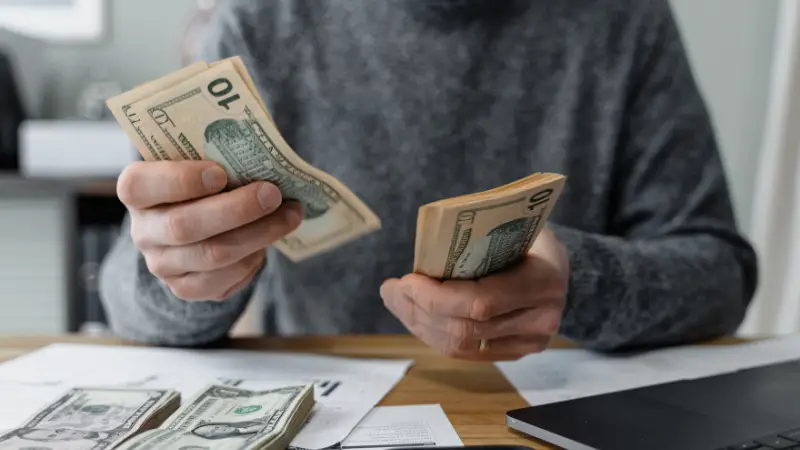 ChatGPT said: A person wearing a gray sweater is sitting at a desk, counting multiple stacks of U.S. $10 bills. The desk is cluttered with paperwork, loose bills, and a laptop, indicating financial activity or budgeting. The image conveys themes of money management, financial planning, or expense tracking.