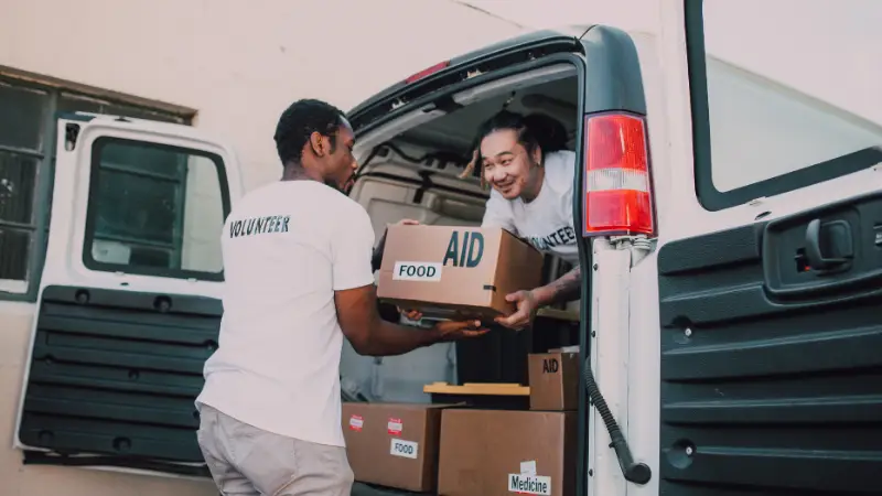 Two volunteers are unloading aid boxes labeled "FOOD" and "MEDICINE" from the back of a white van. One person stands outside the vehicle receiving a box, while the other hands it over from inside. Both are wearing white T-shirts with "VOLUNTEER" printed on the back, highlighting a humanitarian or community service effort focused on delivering essential supplies.