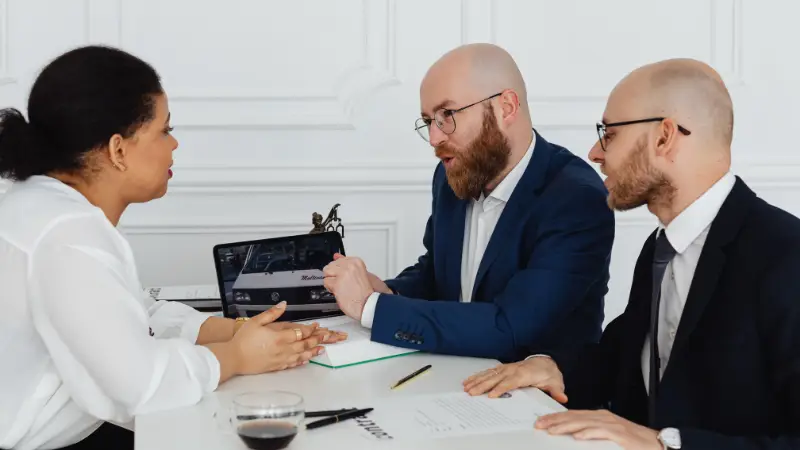 Three professionals are engaged in a serious discussion at a white desk in an office setting. A woman in a white blouse listens attentively as a man in a blue suit gestures toward a laptop screen displaying a vehicle image. Another man in a dark suit sits beside him, observing the conversation. Documents, a pen, and a glass of dark beverage are on the table, suggesting a business or legal consultation.