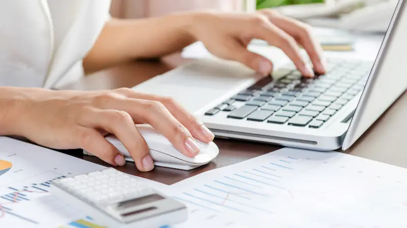 A close-up of a person working on a laptop, using a mouse with one hand while typing with the other. Financial documents with charts and graphs are spread across the desk, along with a calculator, suggesting tasks related to accounting, data analysis, or financial reporting in a professional setting.