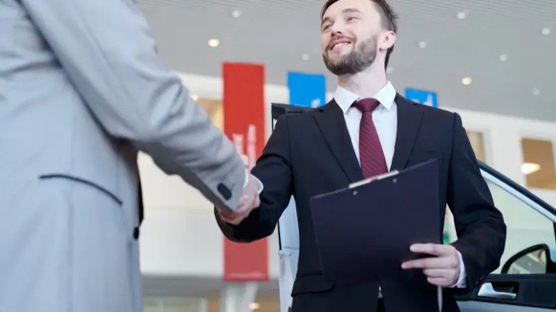 Two men in suits shake hands in a car dealership, with one holding a clipboard and a car visible in the background. Red and blue banners hang on the walls of the modern showroom.