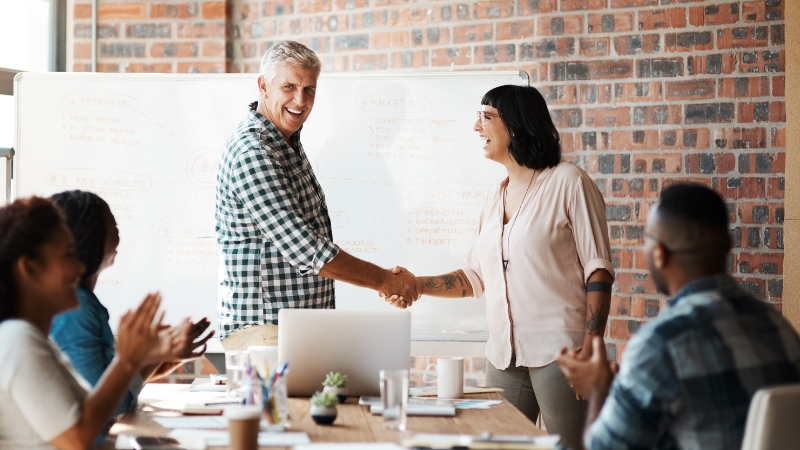 Two people shake hands for accounting services outsourced in a bright meeting room, smiling happily. Others applaud.