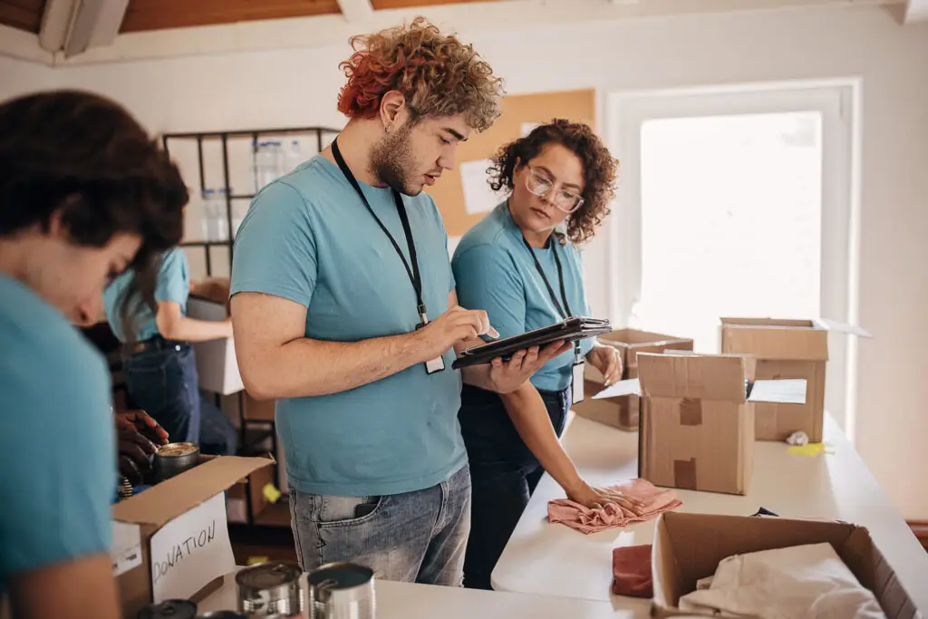 Three volunteers in blue shirts sorting donations in a room. One checks items on a tablet for the 2025 Nonprofit Benchmarking Survey.