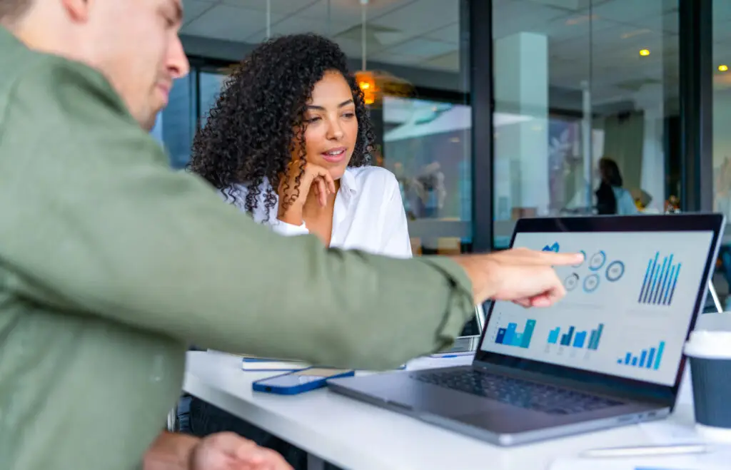 Business man and business woman in a meeting at the office. There is a laptop on the table showing finance charts and graphs. They are looking and pointing at the computer