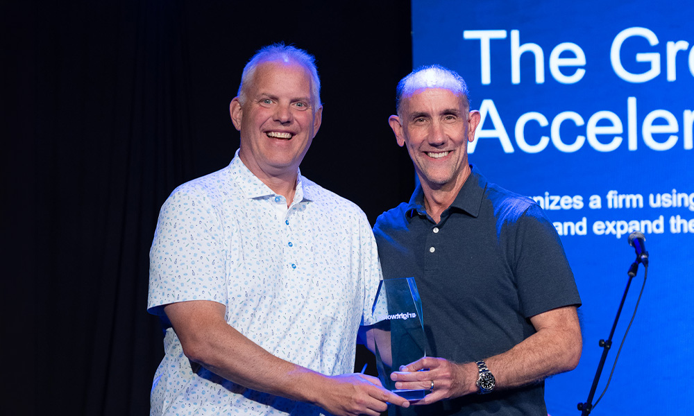 Two smiling men holding a glass trophy on stage in front of a blue digital display screen that reads "The Growth Accelerator Award." The man on the left wears a patterned short-sleeved shirt, and the man on the right wears a dark polo shirt and watch.