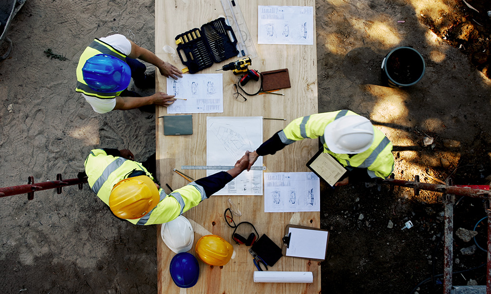 Aerial view of three construction workers in high-visibility vests and hard hats reviewing architectural plans, with two workers shaking hands over the table.
