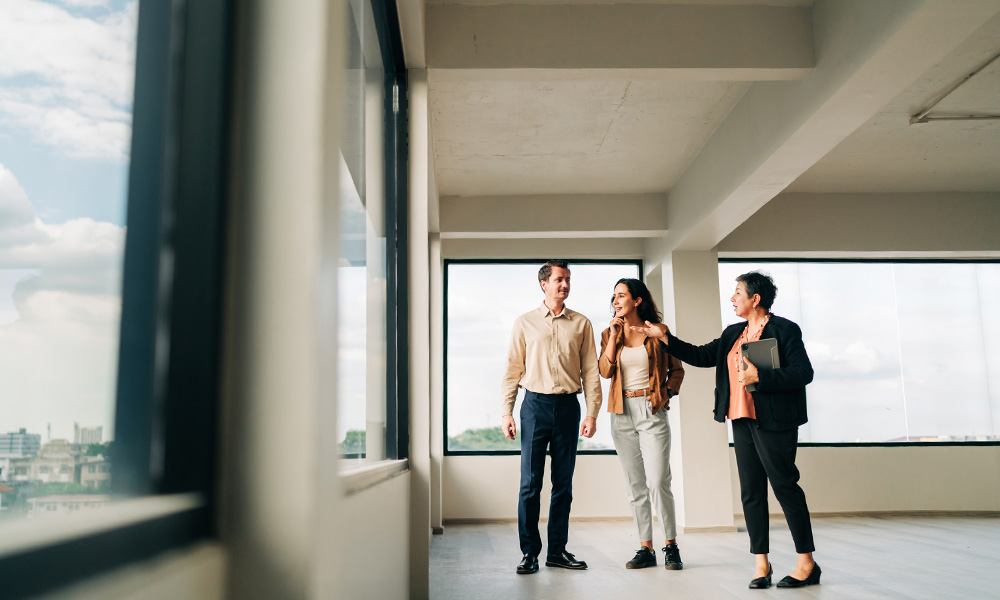 A real estate advisor gestures while showcasing a spacious, empty commercial building interior to a man and a woman.
