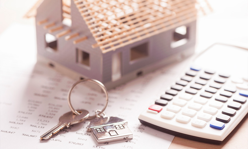 Objects representing real estate finance and investment, including a key, a calculator, and a small, unfinished model house, arranged on financial spreadsheets.